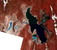 False-color satellite mosaic of an irregular inland lake split by causeways into dark green, blue, and reddish sections amid red mountains and tan flats.