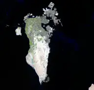 Satellite view of a pale elongated desert island surrounded by black water, with dense gray development along the northeast coast and artificial islands offshore.