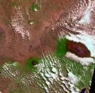 Satellite view of an isolated circular mountain rising from brown plains, with radial valleys, scattered clouds, and a small bright summit ice field.