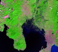 False-color satellite view of a dark crater lake amid radial ridges, pink mudflow channels, and bright green vegetation.