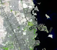 Satellite view of a circular artificial island district with marinas and curved breakwaters, attached to a dense city beside a dark bay.