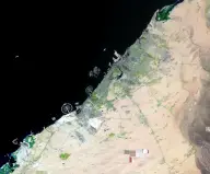 Satellite view of a long pale coastal metropolis beside dark sea and tan desert, crossed by highways and bordered by extensive new development.