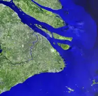 Satellite view of an enormous city surrounding a wide muddy river as it bends toward the sea, with long narrow islands at the estuary.